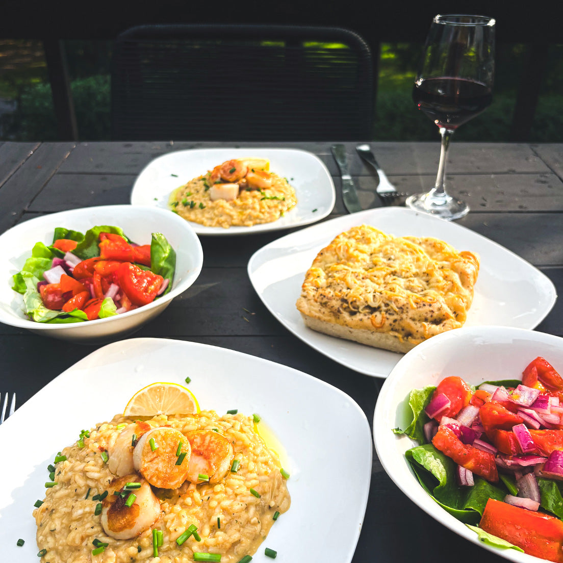 A styled outdoor dining table featuring a seafood risotto with shrimp, a large baked bread or casserole dish, and two fresh salads, alongside a glass of red wine for date night.