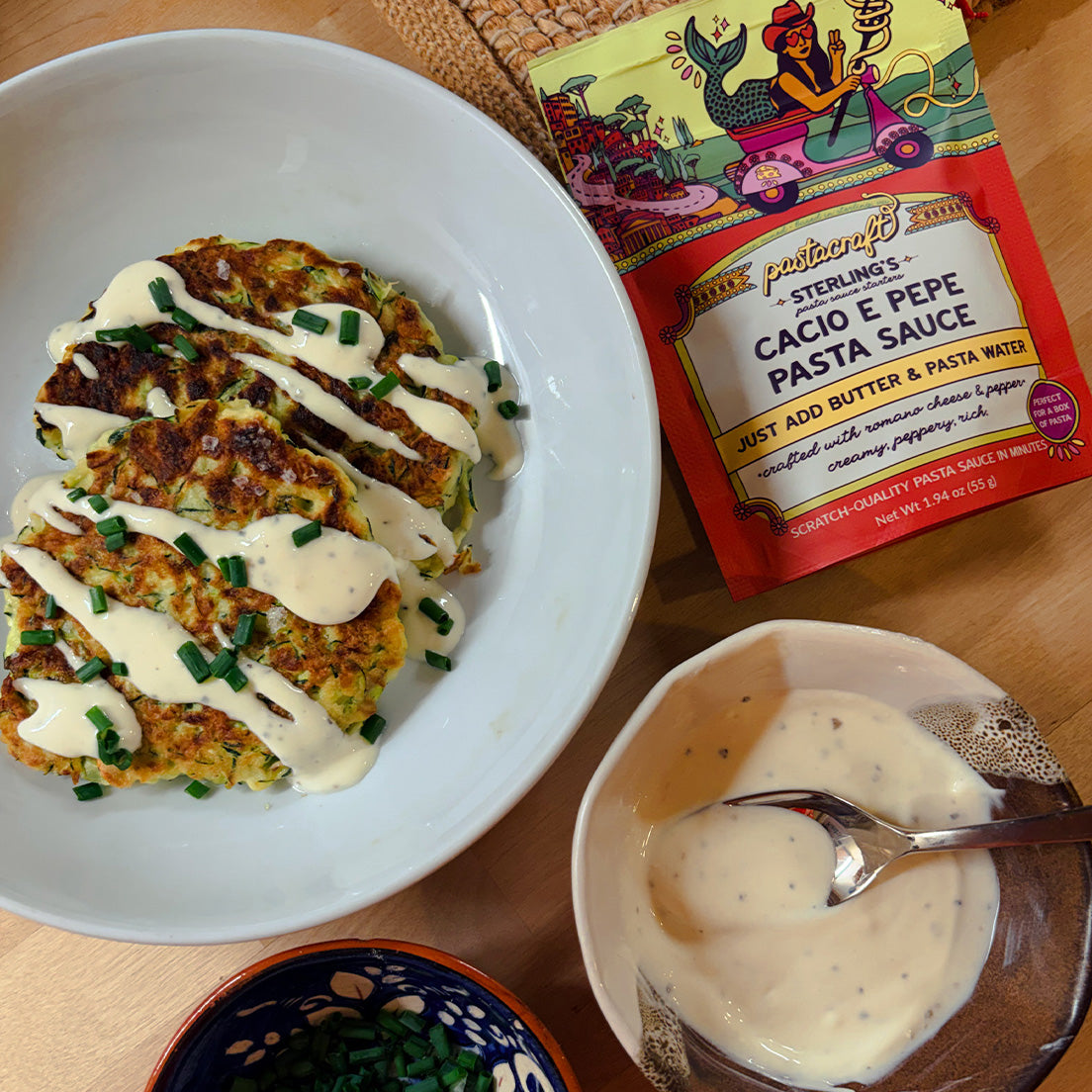 A styled food scene on a wooden table showing a white bowl filled with golden-brown zucchini fritters topped with Cacio e Pepe sauce drizzle and chopped chives, alongside a package of Cacio e Pepe pasta made with Pastacraft pasta sauce Pasta Sauce and a small bowl of the sauce with a spoon.